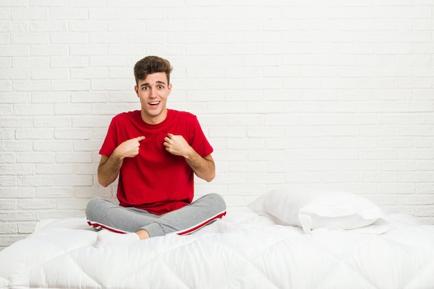 Joven estudiante adolescente hombre en la cama sorprendido señalando con el dedo, sonriendo ampliamente