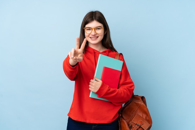 Joven estudiante adolescente sosteniendo una ensalada sobre la pared azul sonriendo y mostrando el signo de la victoria