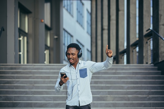 Joven estudiante africano escuchando música desde un teléfono inteligente con auriculares grandes, sonriendo desde la comodidad de usar la aplicación