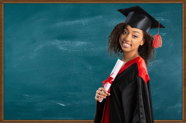 Joven estudiante afro con gorra de graduación y diploma sobre fondo de pizarra