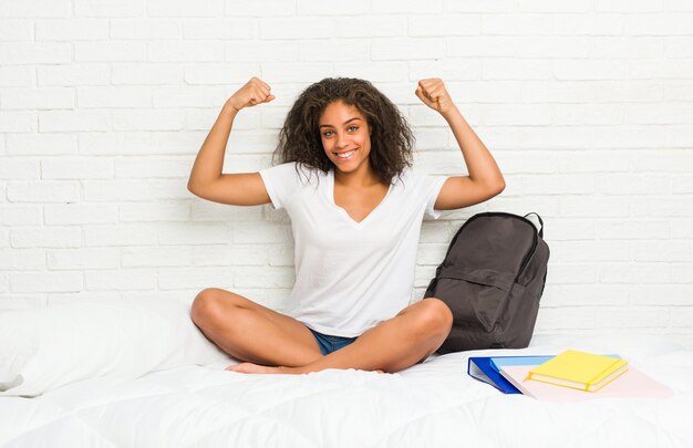 Joven estudiante afroamericana mujer en la cama mostrando fuerza gesto con brazos, símbolo del poder femenino