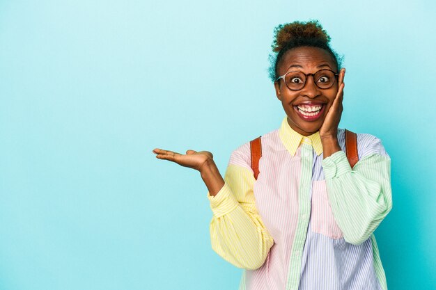 Joven estudiante afroamericana sobre antecedentes aislados tiene espacio de copia en una palma, mantenga la mano sobre la mejilla. Asombrado y encantado.