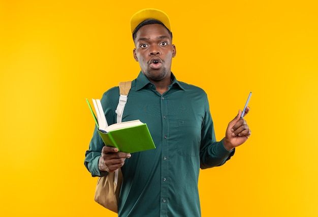 Joven estudiante afroamericano sorprendido con gorra y mochila con libro y bolígrafo