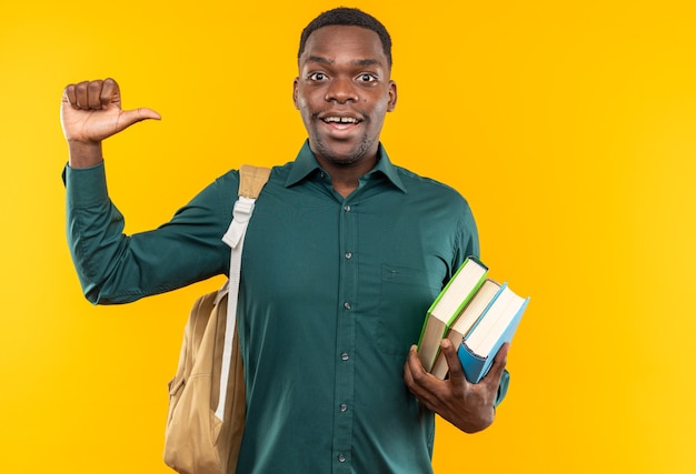 Joven estudiante afroamericano sorprendido con mochila sosteniendo libros y apuntando a un lado