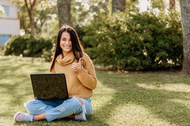 Joven estudiante argentina con un suéter amarillo sonriendo con una computadora portátil en las piernas en un parque