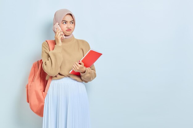 Joven estudiante asiática musulmana molesta con suéter marrón con mochila hablando por teléfono móvil y sosteniendo un libro aislado en fondo blanco concepto de regreso a la escuela