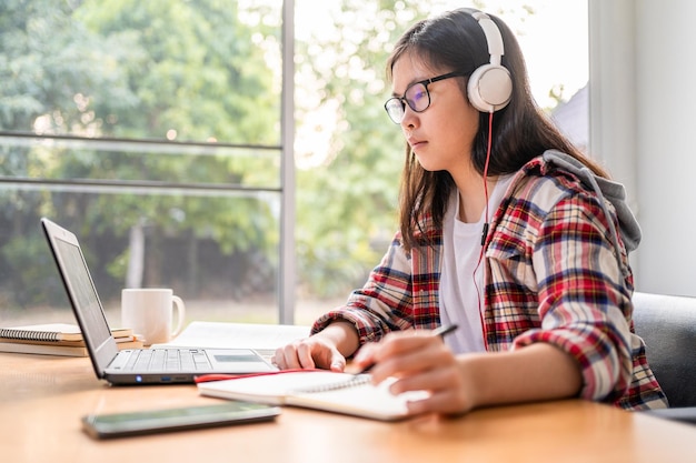 Joven estudiante asiática usando auriculares mientras trabaja y estudia desde casa durante el bloqueo de la ciudad