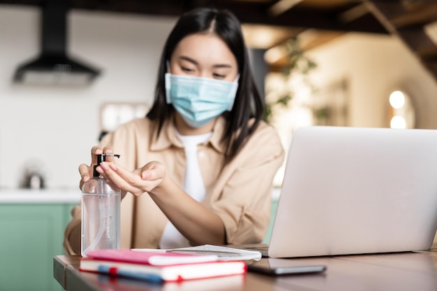 Joven estudiante asiática usando desinfectante de manos en mascarilla médica trabajando en el control remoto de la computadora portátil ...