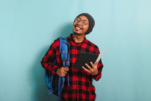 Un joven estudiante asiático que lleva una mochila sostiene libros mientras se enfrenta a un fondo azul