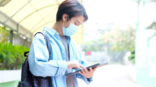 Joven estudiante asiático sosteniendo tableta digital mientras usa máscara de protección