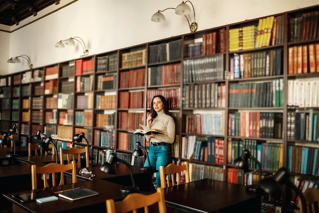 Joven estudiante atractiva leyendo un libro entre las estanterías de la biblioteca