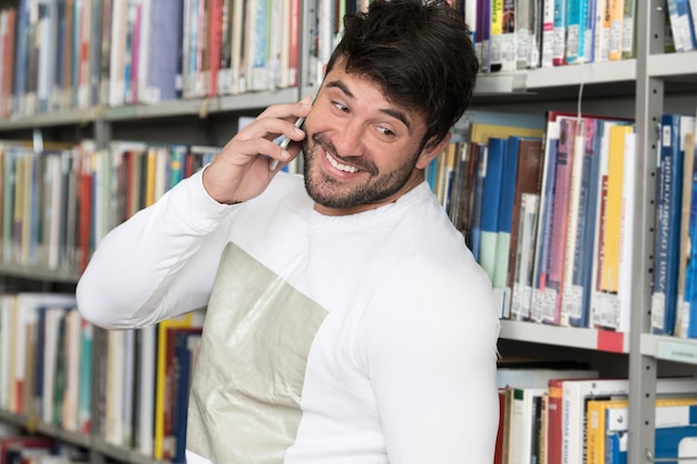 Joven estudiante barbudo hablando por teléfono mientras se prepara para los exámenes en la biblioteca de Univercity