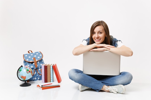 Joven estudiante bastante sonriente casual apoyándose en la computadora portátil y sentado cerca de la mochila del globo, libros escolares aislados