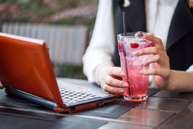 Una joven estudiante bebe limonada fresca mientras trabaja de forma remota en una computadora portátil en un café en un soleado parque de la ciudad