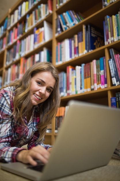 Joven estudiante bonita acostado en el piso de la biblioteca usando la computadora portátil