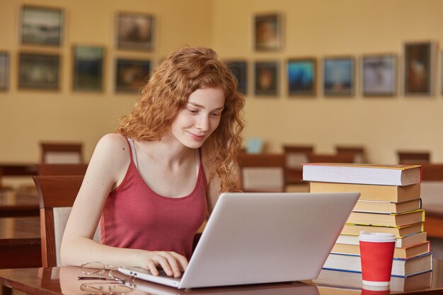 Joven estudiante bonita con laptop y libros trabajando en la biblioteca de la escuela secundaria, sentado en la mesa