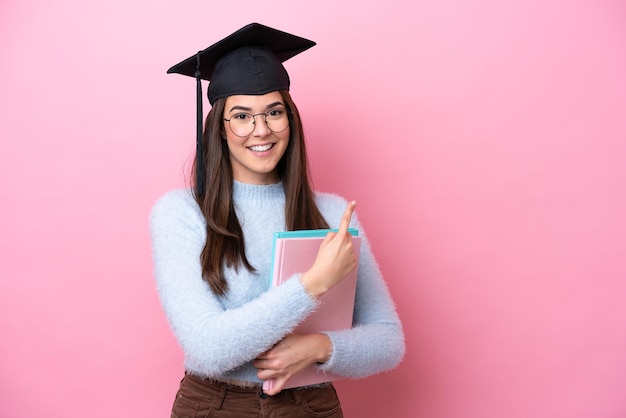 Joven estudiante brasileña con sombrero graduado aislada de fondo rosa apuntando hacia un lado para presentar un producto