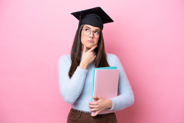 Joven estudiante brasileña con sombrero graduado aislada de fondo rosa con dudas