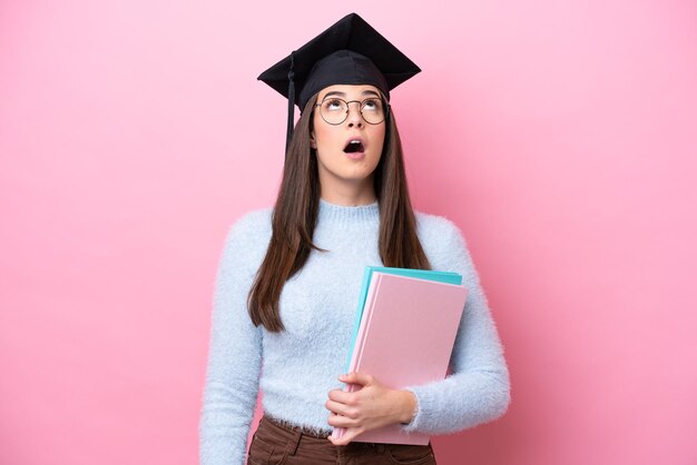 Joven estudiante brasileña con sombrero graduado aislada de fondo rosa mirando hacia arriba y con expresión sorprendida