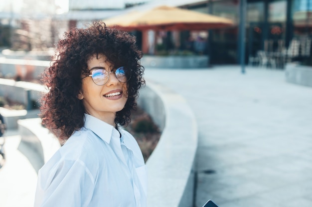 Joven estudiante con cabello rizado y anteojos está sonriendo alegremente mientras mira a la cámara