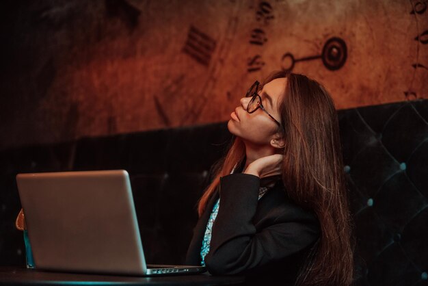 Una joven estudiante cansada que estudia en línea mientras se sienta con una computadora portátil y un cuaderno en la cafetería, una mujer independiente que descansa su cara en la mano con falta de expresión facial mira hacia un lado Enfoque selectivo