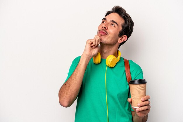 Joven estudiante caucásico bebiendo café aislado de fondo blanco mirando de lado con expresión dudosa y escéptica.