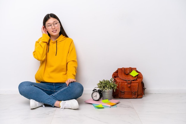 Joven estudiante china sentada en el suelo aislada en una pared blanca pensando en una idea