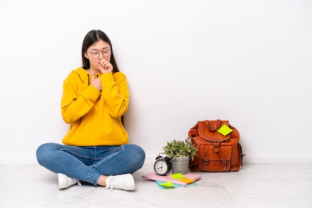 Joven estudiante china sentada en el suelo aislada en una pared blanca sufre de tos y se siente mal