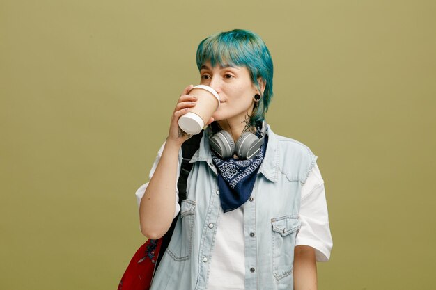 Joven estudiante concentrada usando auriculares y pañuelo en el cuello y la mochila mirando al costado bebiendo café de una taza de café de papel aislada en un fondo verde oliva