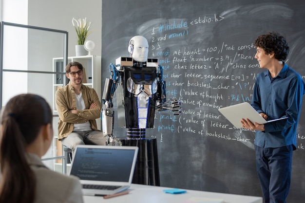 Joven estudiante contemporáneo con portátil de pie junto a la pizarra en el aula y haciendo presentación de robot informático