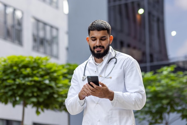 Joven estudiante de doctorado árabe sonriente parado cerca del hospital universitario en uniforme y usando el teléfono