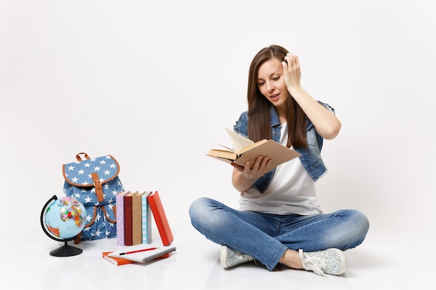 Joven estudiante emocionada en ropa de mezclilla mantenga el libro leído manteniendo la mano cerca de la cabeza sentada cerca del globo, mochila, libros escolares aislados en la pared blanca