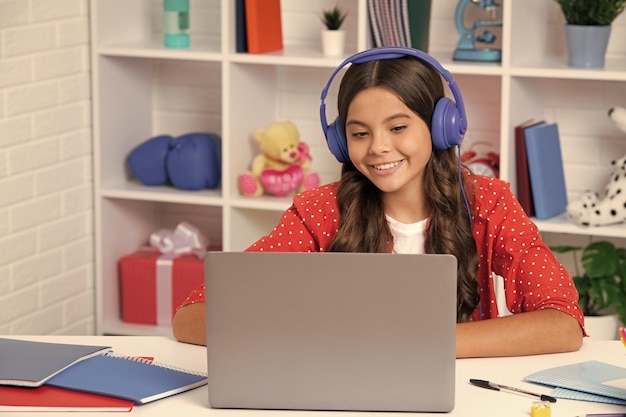 Una joven estudiante de la escuela con auriculares inalámbricos sentada en la mesa usando una computadora portátil cuando estudia