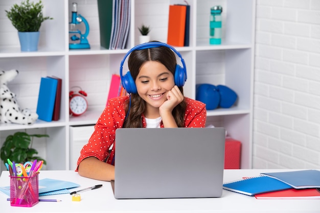 Una joven estudiante de la escuela con auriculares inalámbricos sentada en la mesa usando una computadora portátil cuando estudia