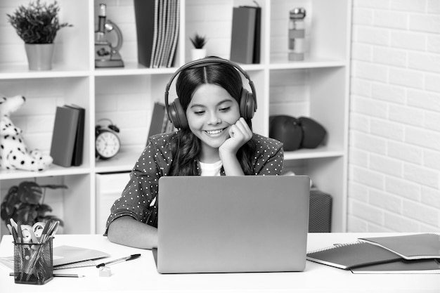 Una joven estudiante de la escuela con auriculares inalámbricos sentada en la mesa usando una computadora portátil cuando