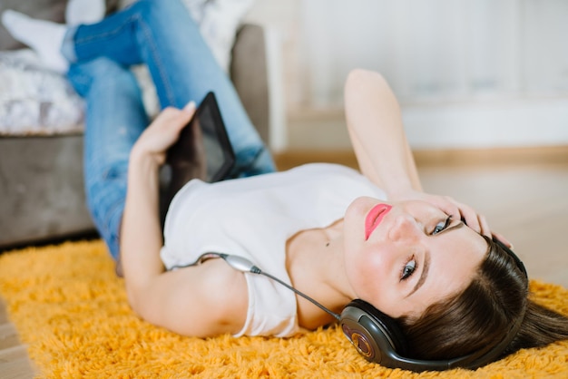 Joven estudiante feliz tirada en el suelo y escuchando música desde una tableta en casa