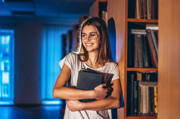 Joven estudiante con gafas en la biblioteca se encuentra cerca de estanterías. La muchacha sonríe y sostiene un libro en sus manos. Preparación para el examen