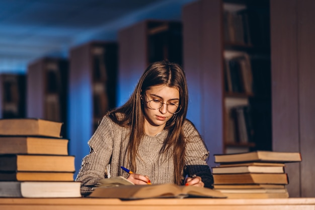 Joven estudiante en gafas de preparación para el examen. Niña en la noche se sienta a una mesa en la biblioteca con una pila de libros