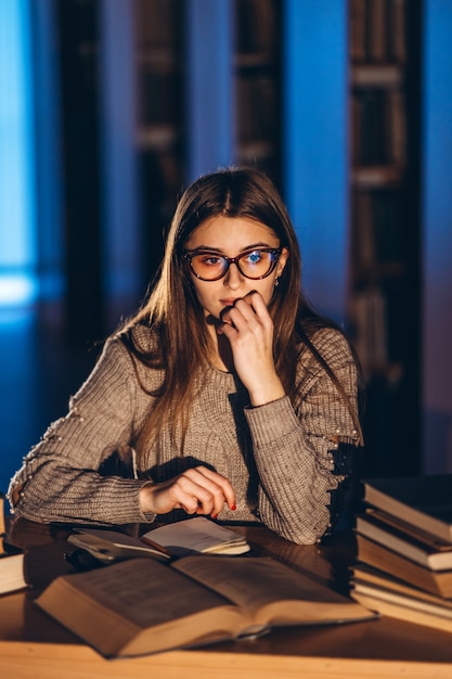 Joven estudiante en gafas de preparación para el examen. Niña en la noche se sienta a una mesa en la biblioteca con una pila de libros