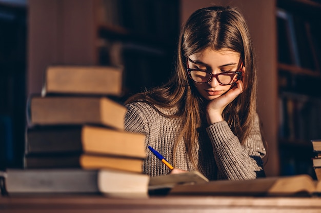 Joven estudiante en gafas de preparación para el examen. Niña en la noche se sienta a una mesa en la biblioteca con una pila de libros