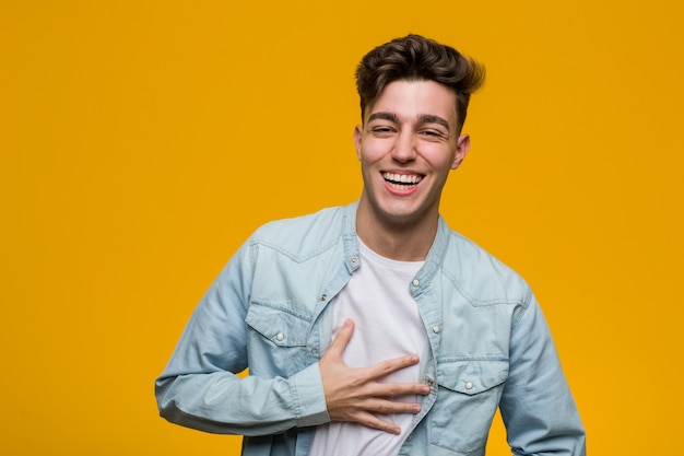 Joven estudiante guapo con una camisa vaquera se ríe alegremente y se divierte manteniendo las manos sobre el estómago.