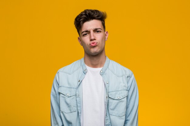 Joven estudiante guapo con una camisa vaquera sopla mejillas, tiene expresión cansada. Concepto de expresión facial