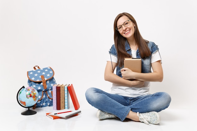 Joven estudiante hermosa alegre en gafas ropa de mezclilla sosteniendo libro sentado cerca del globo, mochila, libros escolares aislados