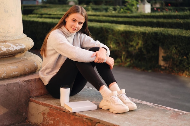 Joven estudiante hermosa sonriente con estilo en un suéter acogedor mirando felizmente a la cámara durante el descanso de estudio al aire libre