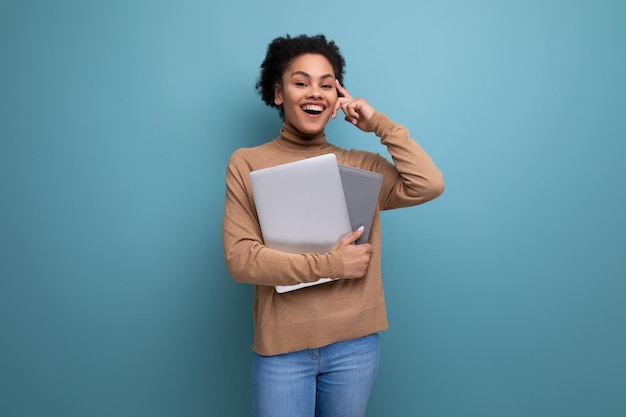 Joven estudiante hispana hermosa con cabello afro rizado en ropa informal sosteniendo una computadora portátil en