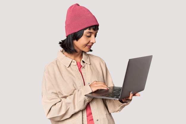 Joven estudiante hispano alegre con computadora portátil y gorra en el estudio