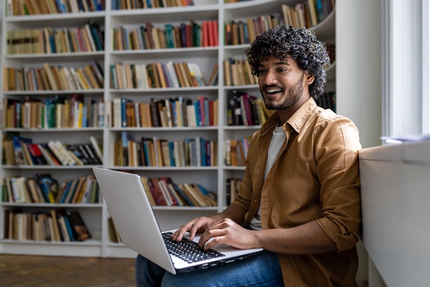 Joven estudiante hispano que estudia dentro de la biblioteca académica entre estanterías hombre escribiendo en la computadora portátil