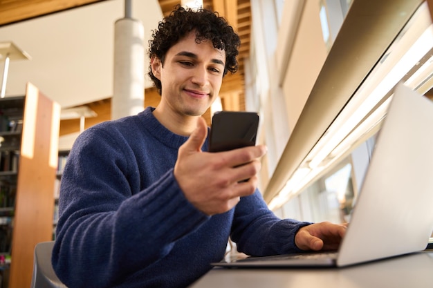 Joven estudiante hispano revisando el teléfono mientras estudia en línea usando una laptop en el campus de la biblioteca
