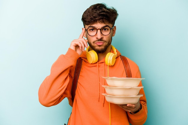 Joven estudiante hispano sosteniendo un tupperware aislado en un fondo azul señalando el templo con el dedo pensando centrado en una tarea