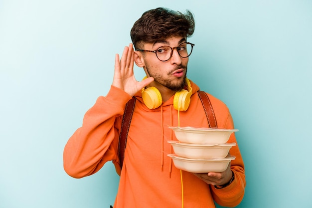 Joven estudiante hispano sosteniendo un tupperware aislado en un fondo azul tratando de escuchar un chisme